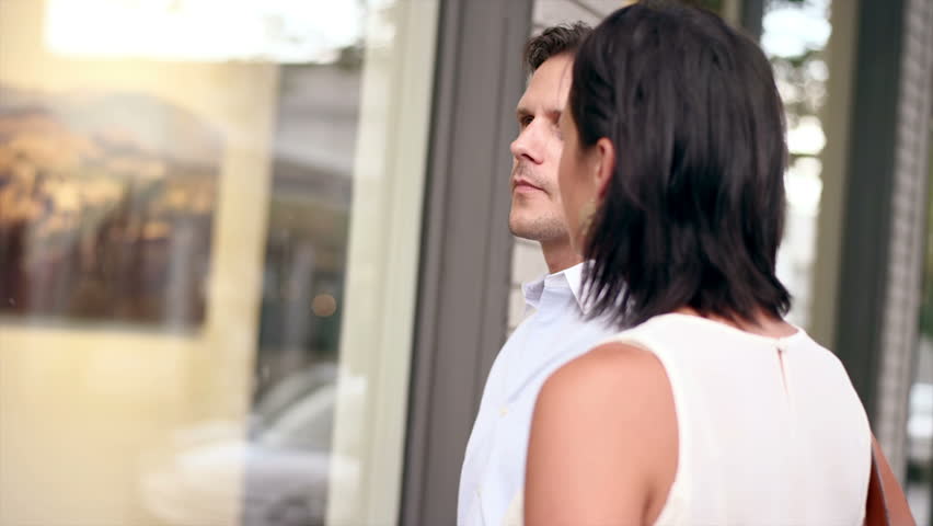 A couple looks through a window and takes some pictures while walking down the street