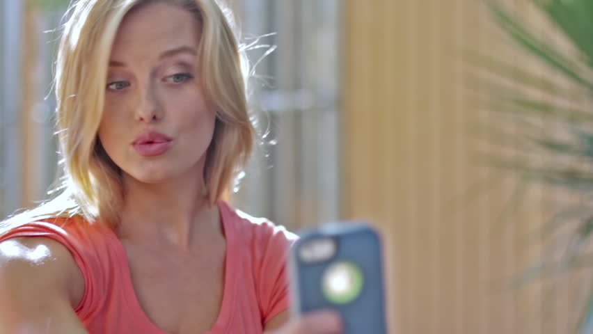Beautiful young blonde woman takes pictures of herself using a smart phone. 
