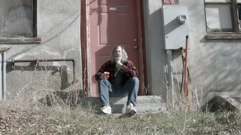 Homeless man broken poor long hair beard HD. Red plaid warm shirt and red Solo cup for drinking. His dynasty is over. Down on luck, poor, hungry and depressed sits on steps of abandoned building.