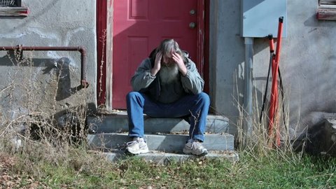 Homeless man cold hungry sad in winter coat HD. Warm winter coat and hood to hide under. Down on luck, poor, hungry and depressed sits on steps of abandoned building. Long hair and big gray beard.