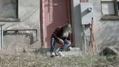 Homeless man broken poor long hair beard HD. Red plaid shirt and red cup for drink and drinking. His dynasty is over. Down on luck, poor, hungry and depressed sits on steps of abandoned building.