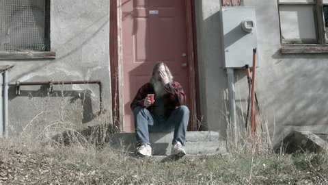 Homeless man sitting on steps sad and poor 2 HD. Red plaid shirt and red cup for drink and drinking. Evicted his dynasty is over. Down on luck, poor, hungry and depressed sits abandoned building.