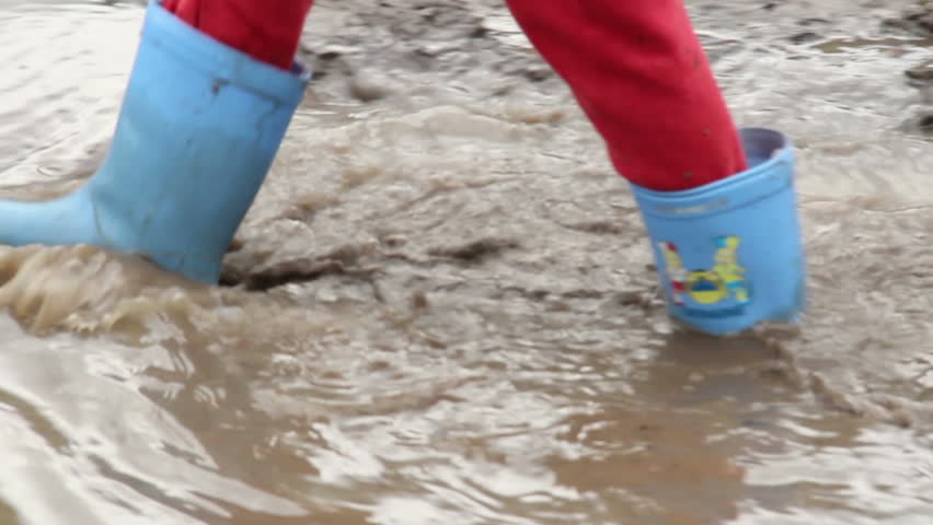 Child feet in rubber boots walk puddle dirt, construction site