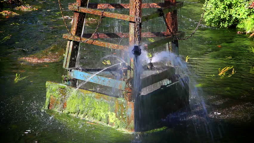 old fountain water mill shooting out water in river with trees sunlight creating shadow close up
