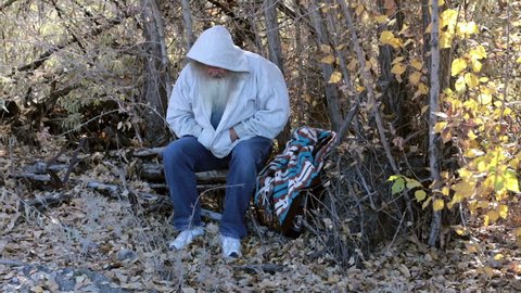 Man homeless in woods wave viewer away HD. Homeless man long hair beard sad and poor. Man down on luck, poor, hungry and depressed sits on tree stump along railroad tracks near urban city.
