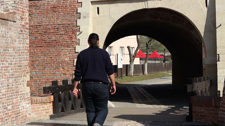 A man enters through the gateway of a medieval fortress, with thick red brick walls and wooden bridge on cure.