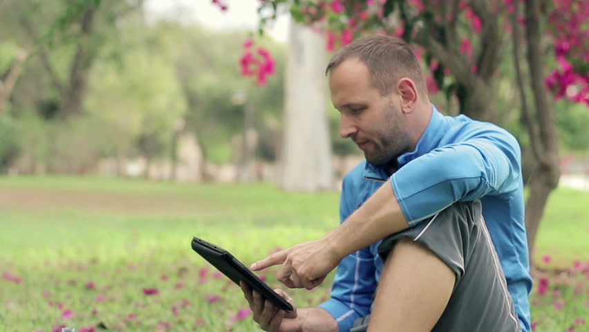 Young sportsman using tablet sitting on grass in the park
