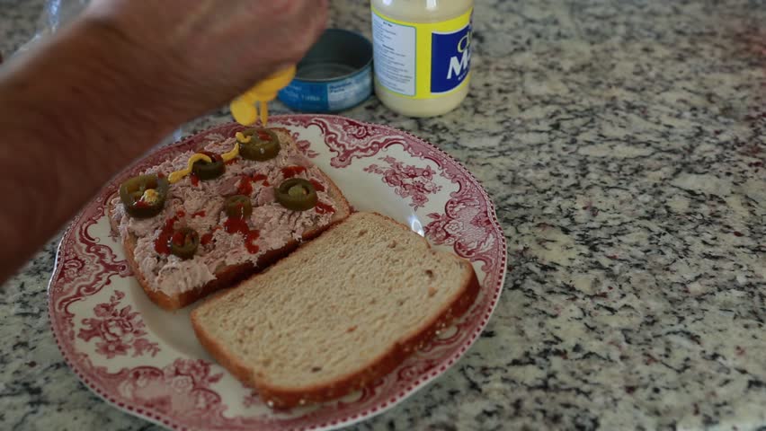 A man making a tuna fish sandwich with jalapeÃ±o's and mustard