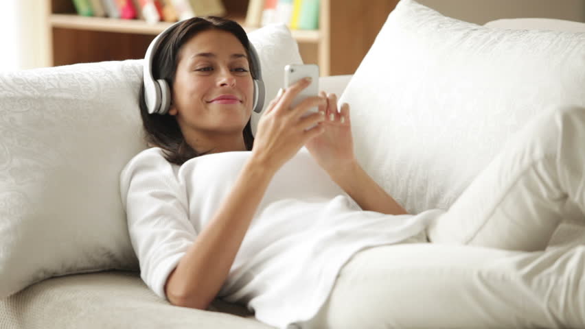 Attractive girl in headphones relaxing on sofa using cellphone looking at camera
