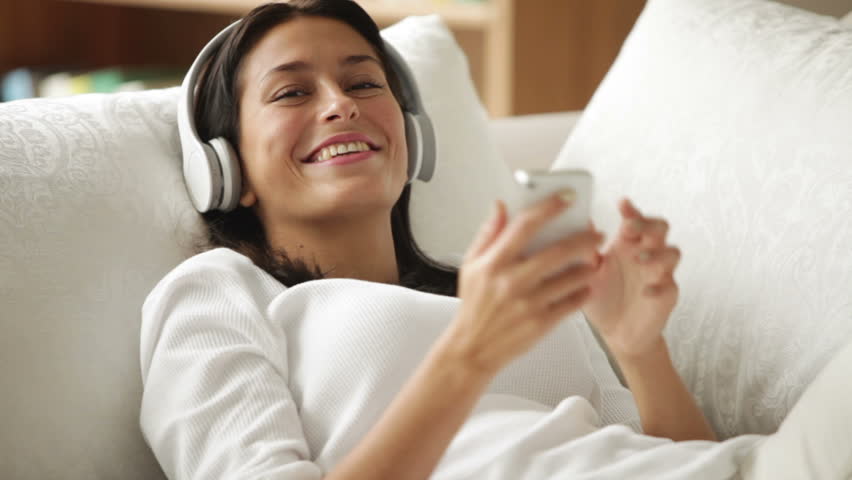 Cheerful girl in headphones lying on sofa using cellphone looking at camera and