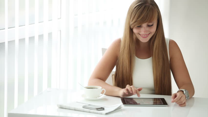 Pretty girl sitting at table with cup of coffee using touchpad looking at camera