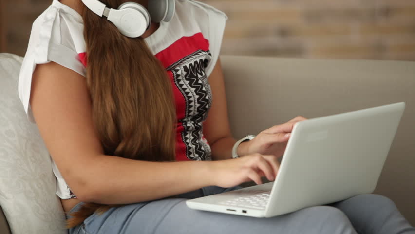 Cute girl in headphones sitting on sofa using laptop looking at camera and