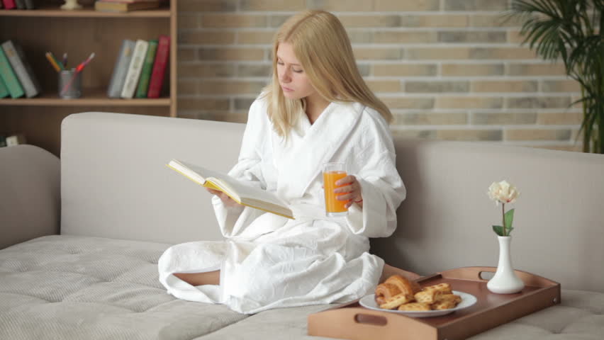 Charming girl in bathrobe sitting on sofa reading book drinkng juice looking at