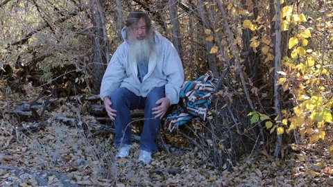 Man homeless in park trees drug addiction itching. HD long hair beard sad and poor. Down on luck, poor, hungry and depressed sits on tree stump along railroad tracks near urban city. Big gray beard.