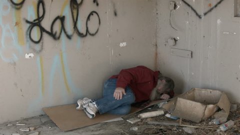 Abandoned building homeless man in corner sleeping HD. Homeless man long hair beard sad and poor sleeping. Man down on luck, poor, hungry and depressed sits on discarded cardboard in urban city.
