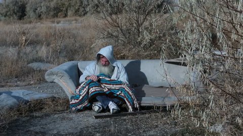 Man outdoor sofa homeless with blanket cold poor. HD long hair beard sad and poor. Man down on luck, poor, hungry and depressed sits on discarded furniture sofa or couch near urban city.