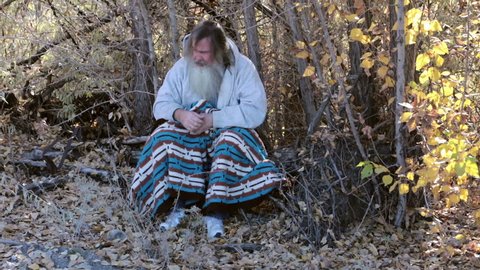 Man homeless and cold with blanket in park trees HD. Homeless man long hair beard sad and poor. Man down on luck, poor, hungry and depressed sits on tree stump along railroad tracks near urban city.