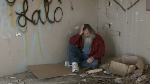 Abandoned building homeless man in corner HD. Homeless man long hair beard sad and poor sleeping. Man down on luck, poor, hungry and depressed sits on discarded cardboard in urban city.