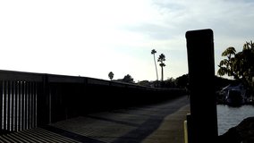 Low angle shot of a generic man in silhouette as he rides a bicycle across a wooden bridge in Newport Beach, California. This clip features the cyclist coming toward and passing the camera.