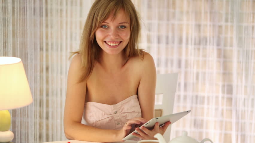 Cheerful blonde girl sitting at cafe using touchpad looking at camera and