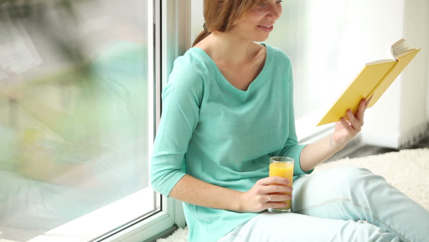 Charming girl sitting by window drinking juice reading book looking at camera