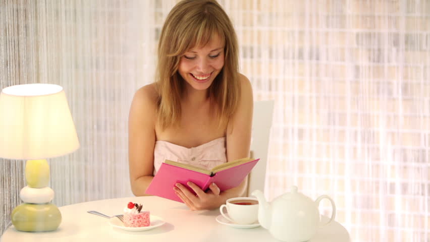 Cheerful girl sitting at cafe reading book looking at camera and smiling