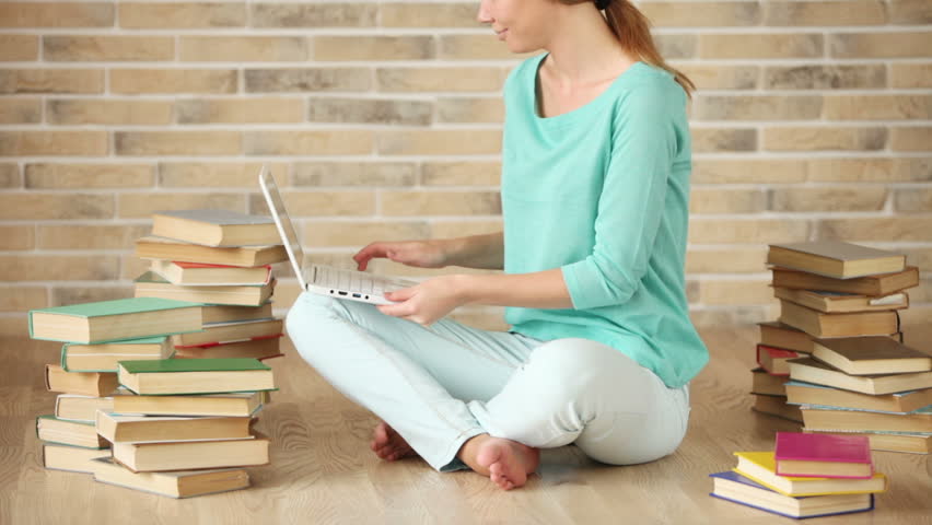 Cheerful girl sitting on floor with books using laptop looking at camera and