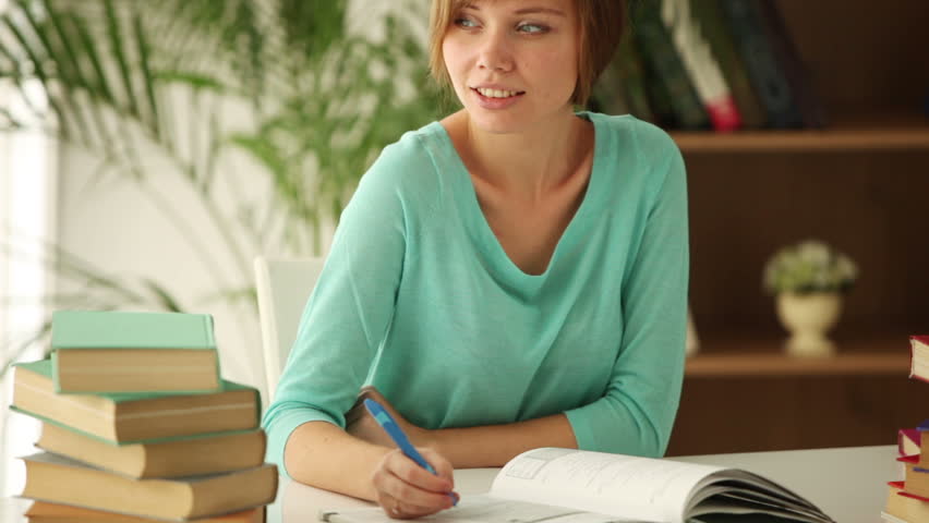 Charming girl sitting at table studying writing in notebook looking at camera