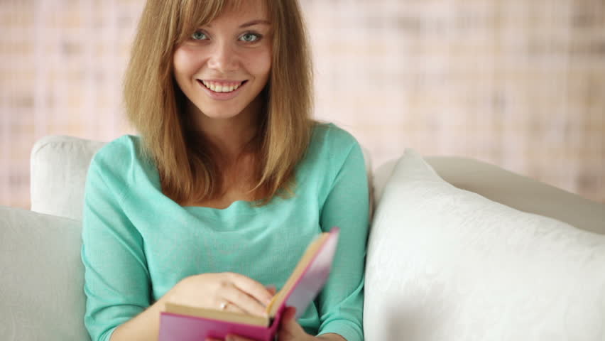 Cheerful girl sitting on couch reading book closing it looking at camera and