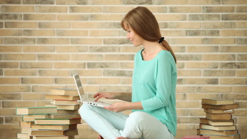 Charming girl sitting on floor with stack of book using laptop looking at camera