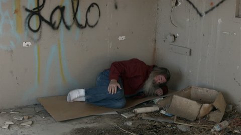 Abandoned building bedroom homeless man in corner HD. Long hair beard sad and poor sleeping. Poor, hungry and depressed sits on discarded cardboard urban city.Needs help to survive. Evicted in cold.