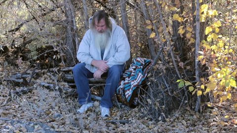 Man homeless hungry and cold in park trees HD. Long hair beard sad and poor. Down on luck, poor, hungry and depressed sits on tree stump along railroad tracks near urban city. Out of work needs help.