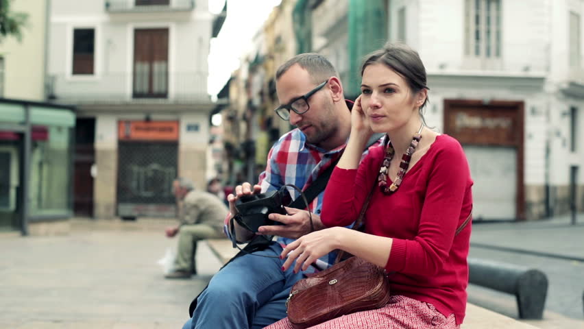 Young couple taking picture with retro camera, sitting on the bench in city
