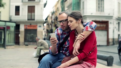 Young couple sitting on bench in the city and watching on smartphone
