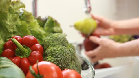 Camera focuses on an assortment of fresh vegetables with a woman washing fruit out of focus in the background.