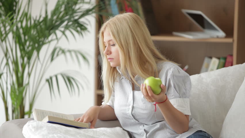 Attractive girl sitting on sofa reading book eating apple and smiling