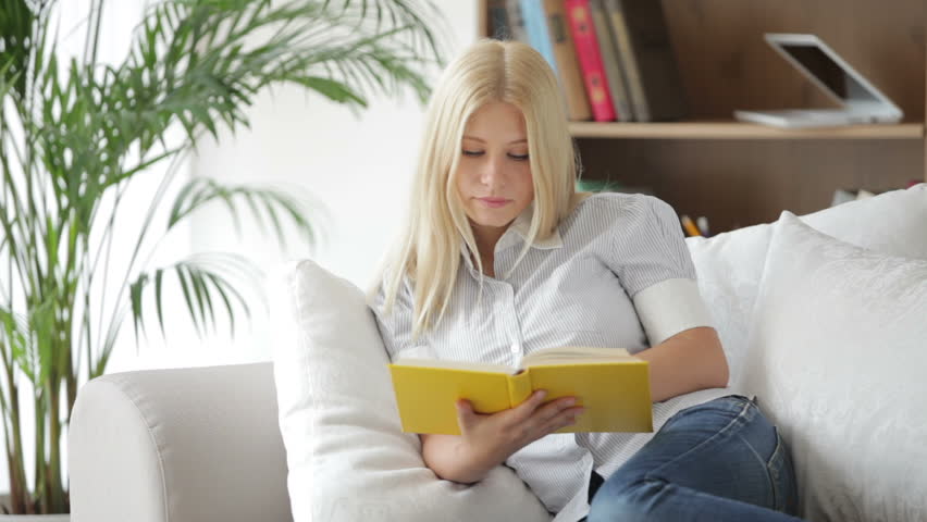 Beautiful girl sitting on sofa reading book looking at camera and smiling