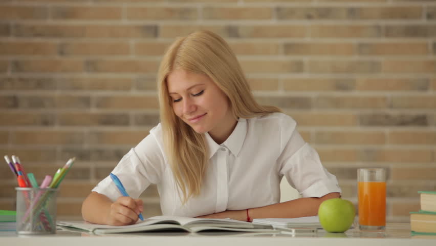 Attractive girl sitting at desk studying writing in notebook looking at camera
