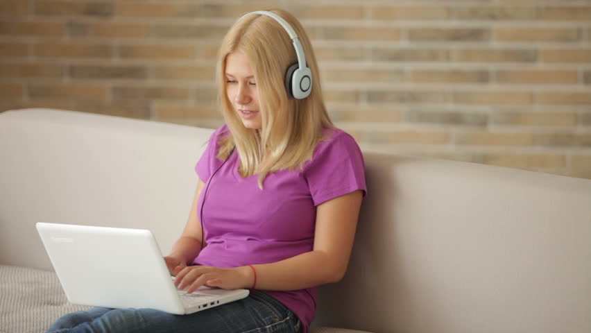 Cute girl in headset sitting on sofa using laptop closing it looking at camera