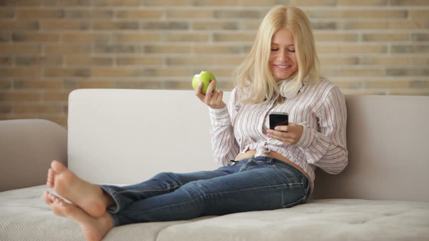 Cute girl in headphones sitting on sofa using cellphone eating apple looking at
