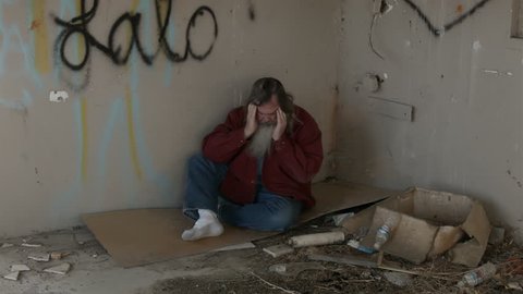 Abandoned building headache homeless man in corner HD. Long hair beard sad and poor sleeping. Hungry and depressed sits on discarded cardboard in urban city. Needs help to survive.
