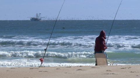A fisherman sets up on the beach.  A commercial fishing boat is on the horizon in the distance.  In 4K UltraHD.