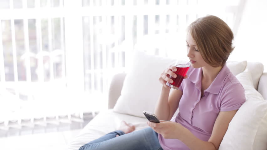 Cheerful girl relaxing on sofa using cellphone drinking juice and smiling at