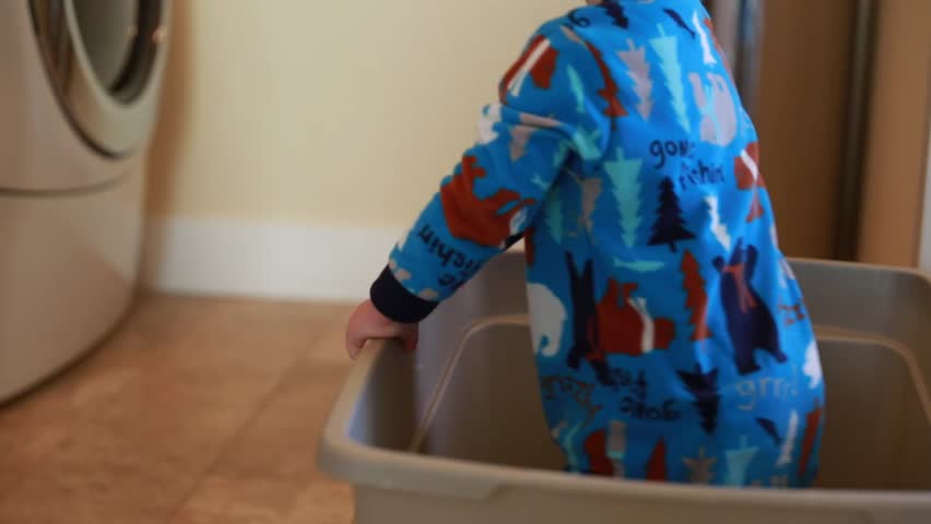 A boy falls over while playing in a laundry basket in his home