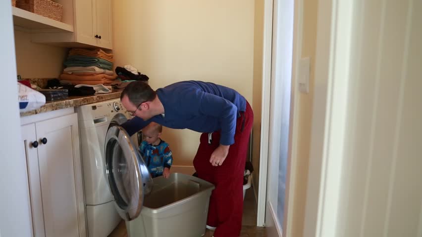 A father does the laundry while his toddler boy plays beside him