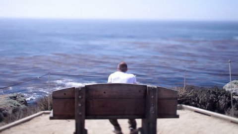 A man takes a seat on a wooden bench at Big Sur, California looks out across the waves