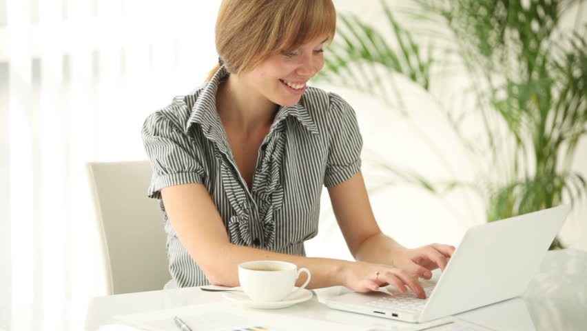 Cute girl sitting at office desk with cup of coffee using laptop looking at