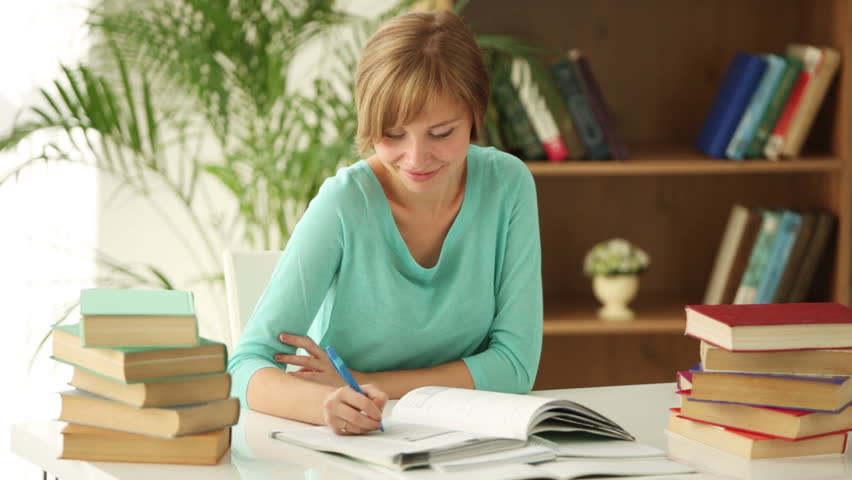 Pretty girl sitting at desk writing in notebook looking at camera and smiling