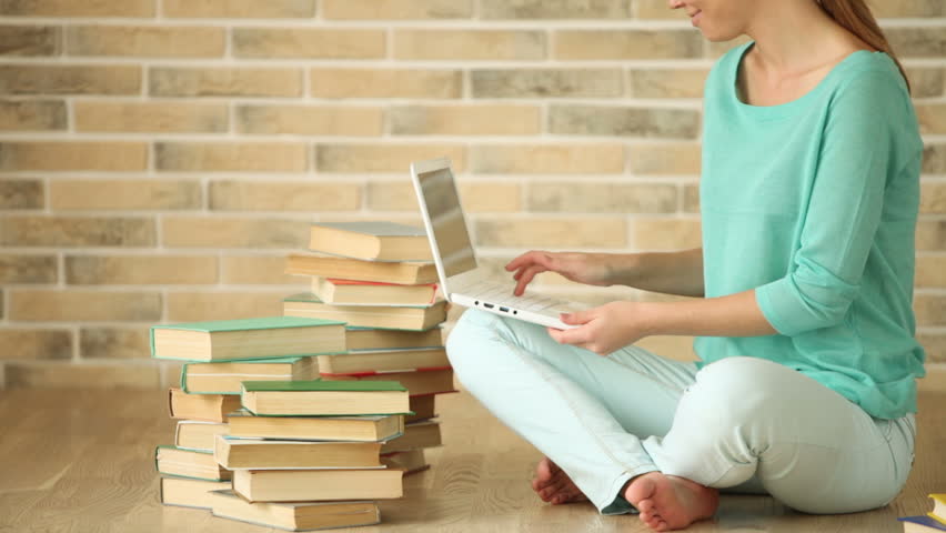 Pretty girl sitting on floor with books using laptop looking at camera and