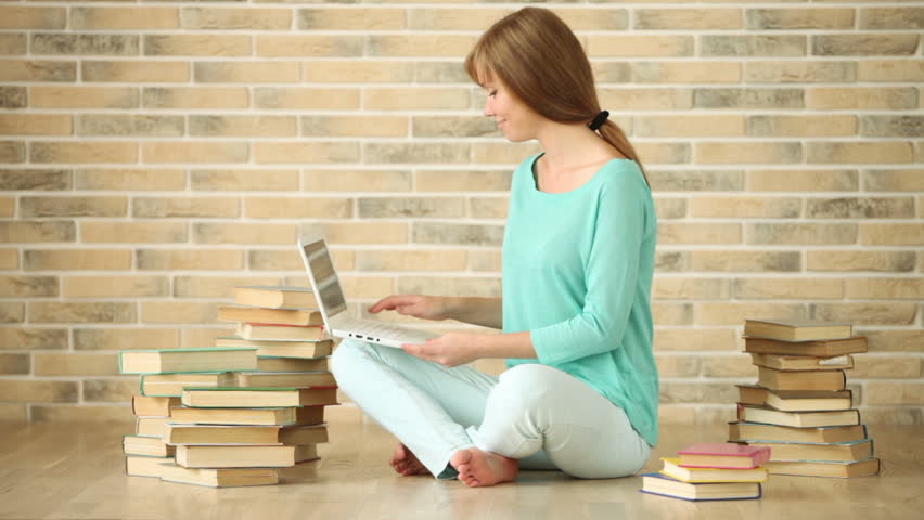 Cute girl sitting on floor with books using laptop looking at camera and smiling